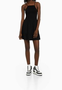Černé šaty s přiléhavým tělem a rozšířenou sukní, se nastavitelnými tenkými ramínky a vrstveným spodním lemem. Nosené s černobílými teniskami.