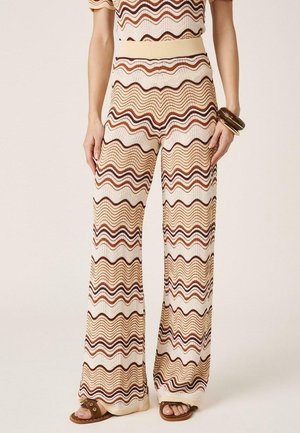 Mujer con pantalones anchos de cintura alta y top a juego con rayas onduladas en beige, marrón y óxido, combinados con sandalias marrones y pulseras.