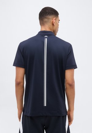 Muž v černé polokošili s krátkými rukávy a bílým vertikálním pruhem na zádech stojí před jednobarevným pozadím.