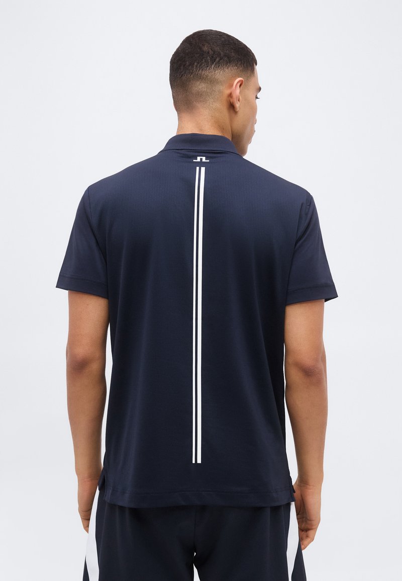 Άνδρας που φοράει μαύρο πόλο μπλουζάκι με κοντά μανίκια και μια λευκή κάθετη ρίγα στην πλάτη, στέκεται μπροστά σε ένα απλό φόντο.