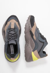 Baskets athlétiques composées d'un mélange de matériaux gris, marron et noir, avec une semelle texturée et un accent jaune vif au talon.