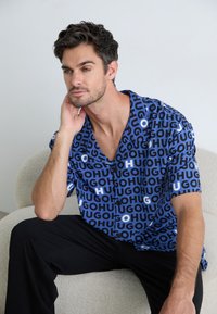 Short-sleeve, blue button-up shirt featuring a bold pattern of "HUG" in black and white. Black pants complement the overall look.