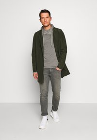 Langer olivgrüner Mantel über einem grauen Kapuzenpullover, kombiniert mit hellgrauen Jeans und weißen Sneakers. Klare Linien und lockere Passform.