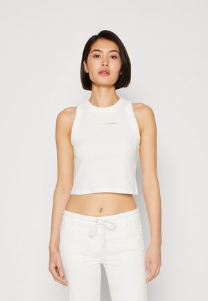 Νεαρή γυναίκα με κοντά μαύρα μαλλιά, φορεμένος λευκό αμάνικο crop top και λευκά παντελόνια με κορδόνι στη μέση, στέκεται μπροστά σε απλό φόντο.