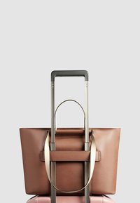 Bruine leren boodschappentas met een vlakke vorm, metalen handvatten en een bevestigde kofferhandgreep, geplaatst op een roze koffer.