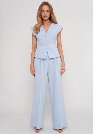 Kobieta ubrana w jasnoniebieską bezrękawnikową kamizelkę z dekoltem w serek z ozdobnymi guzikami oraz pasujące szerokie spodnie, stojąca na jednolitym tle.