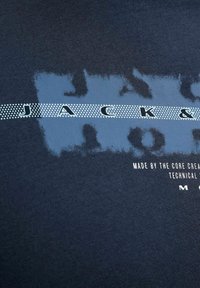 Tissu bleu foncé avec « JACK & » en lettres blanches en pointillés sur un fond rectangulaire bleu délavé et texte blanc plus petit en dessous.