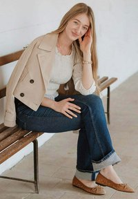 Gabardina beige, blusa blanca con textura, jeans de mezclilla azul oscuro y bailarinas marrón claro con tachuelas doradas. Banco de madera contra una pared clara.