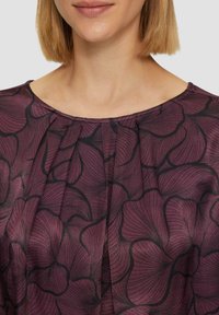 Paarse blouse met zwart bloemenpatroon, geplooide halslijn en lichte, gladde stof. Heeft een ronde kraag en korte mouwen.