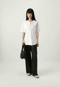 Woman with long black hair wears loose white top and wide black pants, holding a black woven handbag, standing against plain background.