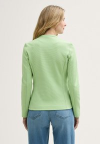 Veste vert clair à manches longues, au design ajusté et au tissu texturé. La vue de dos montre des lignes épurées et un style minimaliste. Associée à un jean bleu.