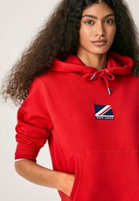 Felpa rossa con cappuccio, tasca frontale, cappuccio con cordino e logo applicato. Presenta dettagli a coste sui polsini e una texture di tessuto liscia.