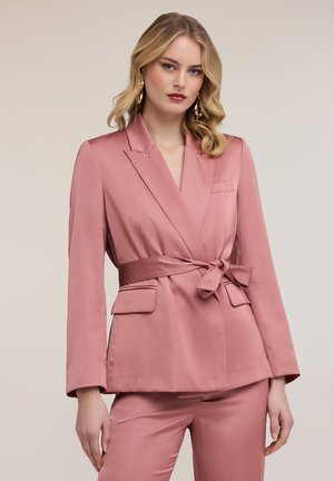 Kvinde med bølget blondt hår iført en pink satinbælte blazer og matchende bukser, stående foran en neutral baggrund.