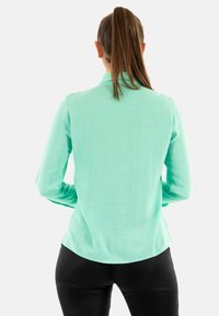 Camicia a maniche lunghe verde menta con una texture liscia, con colletto e orlo curvato, abbinata a pantaloni neri aderenti, vista da dietro.