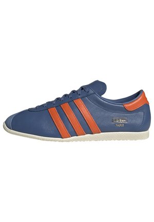 Zapatilla Adidas baja de color azul con rayas naranjas y lengüeta en el talón, suela blanca y marca "Adidas Paris" en el lateral.