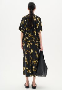 Abito nero con stampa floreale, dal taglio morbido, maniche corte e gonna svasata. La modella tiene una grande borsa in pelle nera, indossando sandali neri con zeppa.