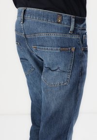 Dark blue denim jeans with a straight-leg design, featuring dual back pockets, contrasting stitching, and a brand tag on the waistband.