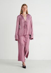 Completo in seta composto da blusa e gonna in malva con dettagli in pizzo, presenta una chiusura con lacci frontale e maniche lunghe, completato da una gonna con spacco laterale.