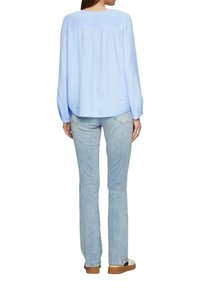 Blouse bleu clair à manches longues en tissu texturé, coupe ample, avec détail froncé à l'arrière, associée à un jean en denim clair et des chaussures décontractées.