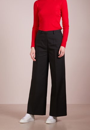 Person trägt ein rotes langärmeliges Oberteil, hoch taillierte schwarze weite Hosen und weiße Turnschuhe und steht auf einem schlichten Boden.