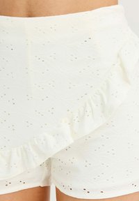 Pantalones cortos blancos con una cintura con volantes, tela texturizada con un patrón de ojales florales y pequeños cortes a lo largo.