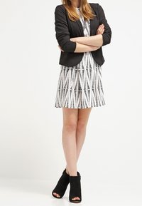 Musta bleiseri valkoisen mekon päällä, jossa on musta geometrinen kuvio, yhdistettynä mustiin avonaisiin nilkkabootseihin. Ominaisuuksiin kuuluu tiukka muotoilu ja hihaelementit.