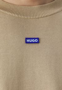 Beige neule, jossa on ribbauskaulus, esittelee sinisen suorakulmaisen tägin, jossa on valkoiset "HUGO"-tekstit, korostaen neulepintaa ja modernia muotoilua.