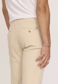 Pantalons beiges avec un fini texturé, dotés de poches arrière avec fermetures à bouton et d'une petite étiquette sur la taille.