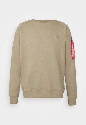 Beżowa bluza z długim rękawem i okrągłym dekoltem, z małą naszywką na piersi oraz czerwonym żółtym oznaczeniem "Remove Before Flight" na kieszeni z suwakiem na lewym rękawie.