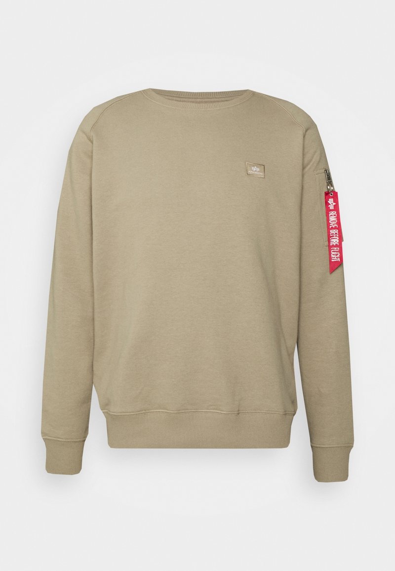 Sweat-shirt beige à manches longues avec col ras du cou, petit écusson sur la poitrine et étiquette rouge "Remove Before Flight" sur la poche zippée de la manche gauche.