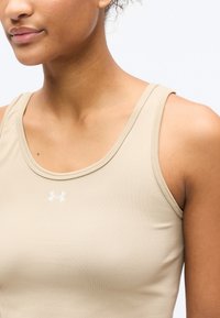 Κοντινό πλάνο ενός ατόμου που φοράει μπεζ αθλητικό τοπ χωρίς μανίκια με μικρό λευκό λογότυπο Under Armour στο στήθος.