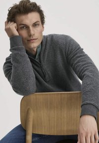Grauer Fleece-Pullover mit Reißverschluss und strukturiertem Stoff, kombiniert mit einem dunkelblauen Hemd mit Knöpfen, vor einem hellen Hintergrund mit einem Holzstuhl.
