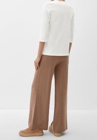 Long brown wide-leg pants in soft knit fabric paired with a fitted white three-quarter sleeve top, viewed from the back with beige sneakers.