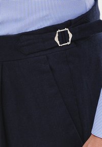 Ciemnoniebieskie spodnie z teksturowanej tkaniny, z szerokim pasem w talii i metalowym zapięciem. Widoczna koszula w kratę powyżej.