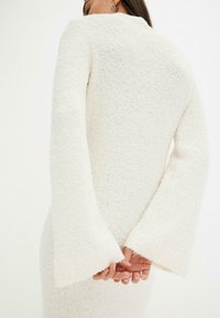Abito bianco in maglia texturizzata con scollatura alta e maniche svasate, che mette in mostra un materiale caldo e una silhouette aderente.