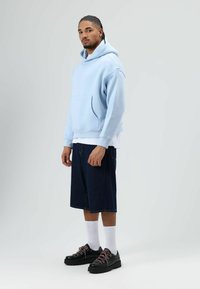 Sweat à capuche bleu clair avec une poche avant, associé à un short en denim foncé et des chaussures noires avec des lacets roses. Chaussettes blanches visibles.