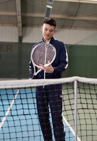 Tennisracket met een wijd, wit snaarpatroon vastgehouden door een persoon in een blauwe trainingsoutfit, staande bij een tennisnet op een baan.