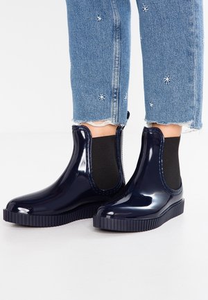 Bottes de pluie noires brillantes à la cheville avec panneaux latéraux élastiques, portées avec un jean denim bleu présentant de petits motifs d'étoiles blanches.