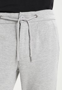 Pantalones de chándal grises con cintura ajustable, que cuentan con un cierre de botón, textura suave y bolsillos laterales. Los cordones tienen puntas de metal.