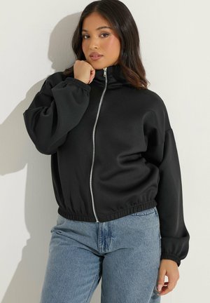 Jonge vrouw poseert in een zwarte jas met rits en blauwe spijkerbroek tegen een effen lichte achtergrond met zachte schaduw.