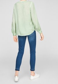 Blusa verde chiaro, semi-trasparente, con maniche lunghe e polsini elasticizzati, abbinata a jeans skinny blu e sneakers bianche. Texture liscia e orlo curvo.