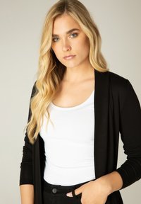 Zwarte lange mouwen cardigan over een witte tanktop, met een relaxte pasvorm en een zachte textuur. Model poseert met een hand in de zak.