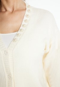 Krémový cardigan vyrobený z měkkého materiálu s V-výstřihem, zdobený perlovo-ovitými akcenty podél límce a klasickým zapínáním na knoflíky.