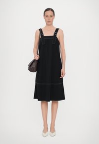 Rochie neagră fără mâneci, cu bretele late, având cusături albe. Formă A-line și material fluid. Modelul ține o geantă mică maro și poartă pantofi cu toc bej.