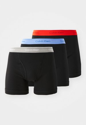 Trois paires de slips boxers noirs avec des ceintures élastiques en rouge, bleu et gris, portant le logo "Calvin Klein" sur chaque bande.