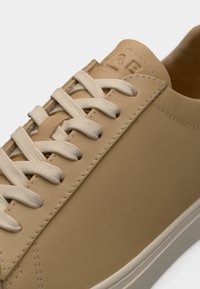 Κοντινό πλάνο μπεζ αθλητικού παπουτσιού με εκρού κορδόνια, εμφανές ράψιμο και ανάγλυφα αρχικά της μάρκας στη γλώσσα.