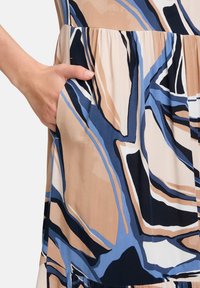 Gemustertes Kleid mit abstrakten Formen in Beige, Marineblau und Blautönen. Der Stoff ist glatt; das Kleid verfügt über eine Seitentasche und hat ein geschichtetes Design am Saum.