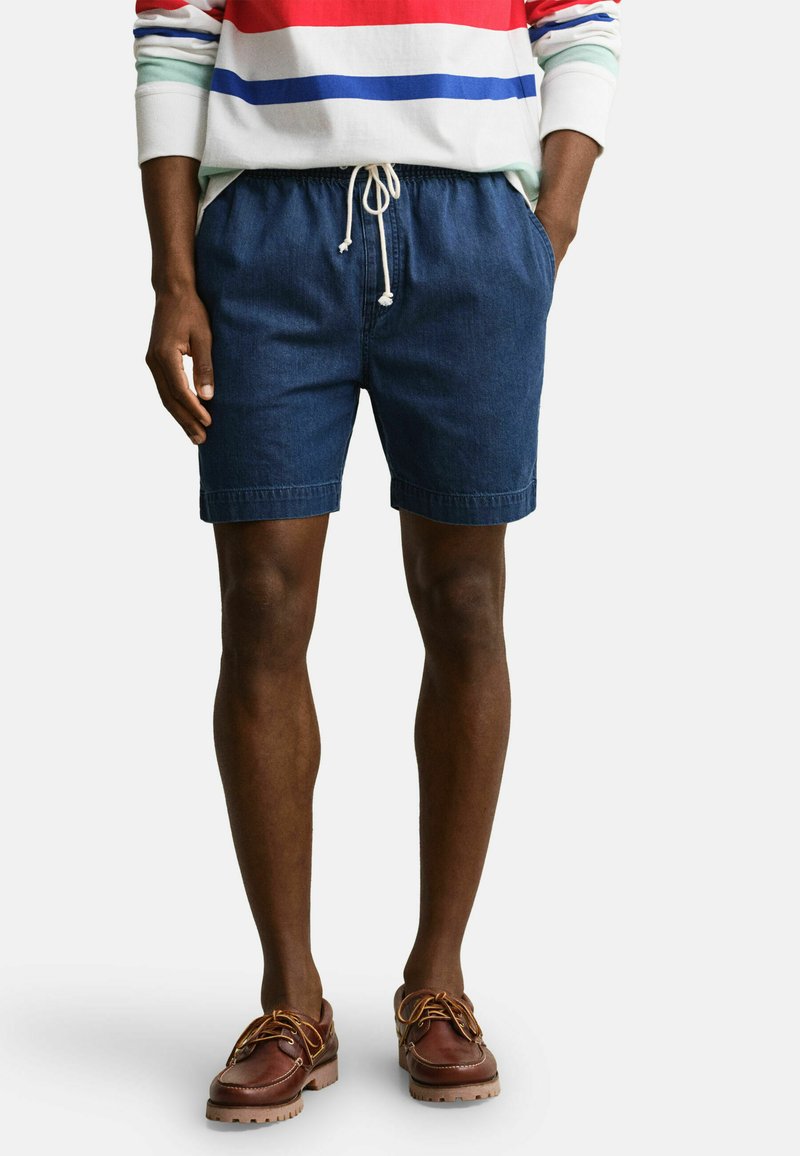 Uomo che indossa pantaloncini blu scuro con coulisse, maglietta a maniche lunghe a righe multicolori e scarpe da barca in pelle marrone, in piedi con le mani nelle tasche.