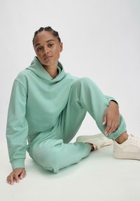 Minzgrüner, übergroßer Hoodie und passende Jogginghosen aus weichem Stoff. Glatte Textur, lässige Passform und elastische Bündchen. Beigefarbene Sneakers runden das Outfit ab.