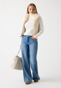Gilet in pelliccia sintetica beige sopra un dolcevita bianco, abbinato a jeans a gamba larga blu chiaro e una borsa grigia. Texture morbide e tonalità neutre.